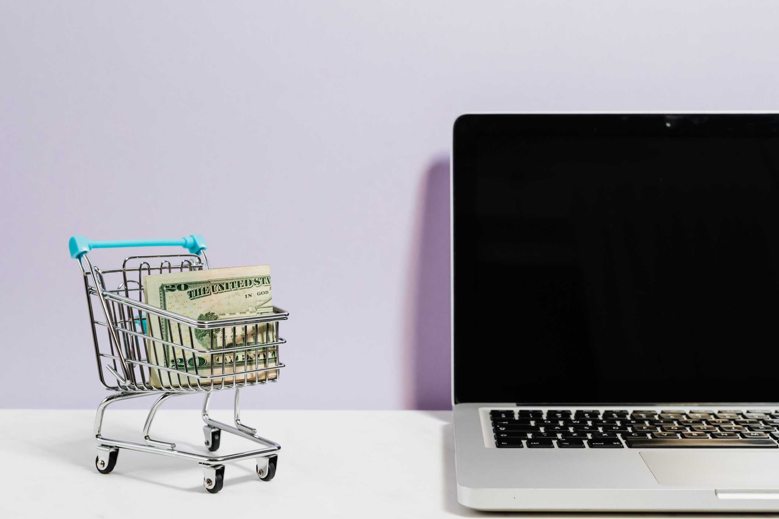 A shopping cart containing US dollar banknotes next to a laptop