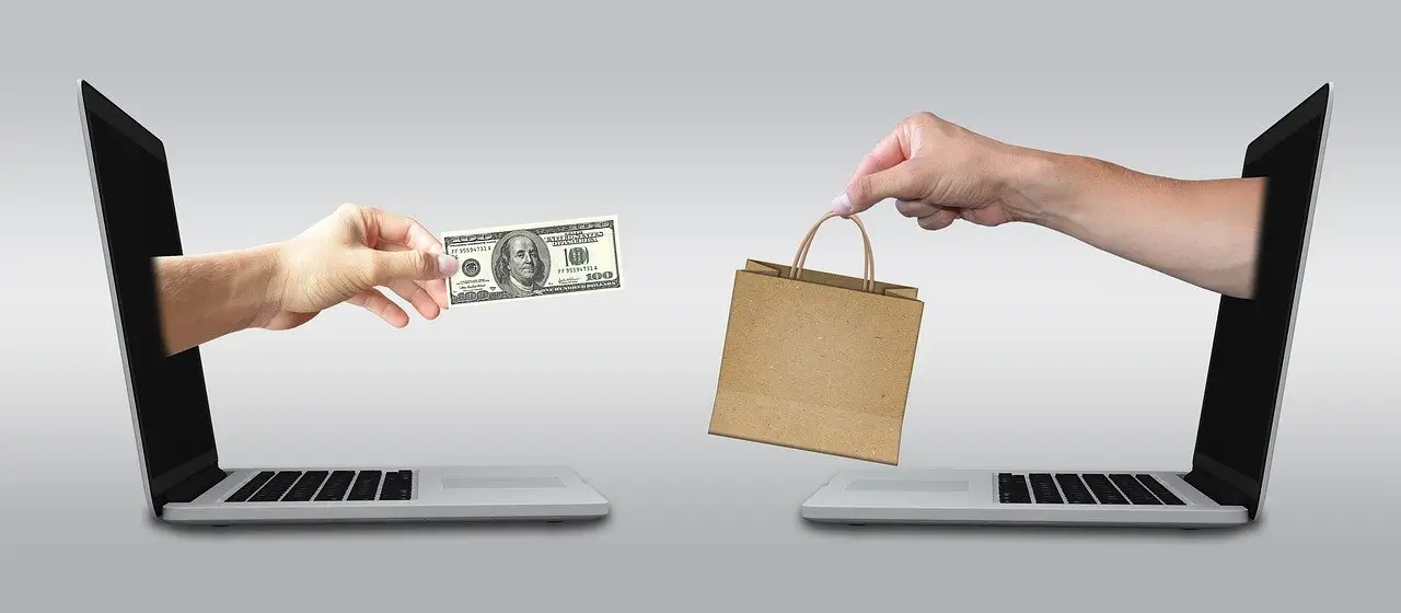 A hand extending from two laptops exchanging money for a bag.